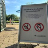 An jedem der drei Freibäder in Recklinghausen hat die Stadt Recklinghausen diese Klappaufsteller aufgestellt, auf denen steht: Bitte halten Sie Ihre Taschen zur Kontrolle bereit! Dazu befinden sich zwei Verbotsschilder auf dem Aufsteller: Eins zeigt durchgestrichen ein zerbrochenes Glas und eine zerbrochene Flasche. Das zweite zeigt durchgestrichen eine Pistole, ein Pfefferspray, ein Klappmesser und einen Teleskopschlagstock. Im Hintergrund ist der Eingangsbereich des Südbads samt Drehkreuz zu e