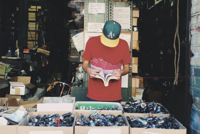 Ein Mann blättert auf einem Flohmarkt in einem Buch. Vor ihm stehen Kisten mit gebrauchten Kleinigkeiten.
