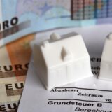 Symbolfoto: Spielzeughäuschen stehen auf Geldscheinen und einem Steuerbescheid.