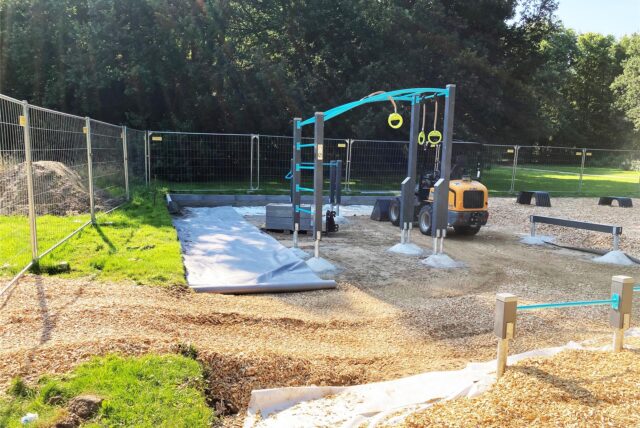Blick auf die Baustelle, auf der der neue Fitnessparcours im Backumer Tal in Herten entsteht. Zwischen den Sportgeräten steht ein kleiner Bagger.