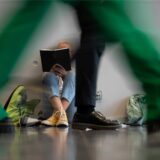 Eine Frau sitzt auf dem Boden und ist in ein Buch vertieft. Währenddessen gehen Menschen schnellen Schrittes vorbei.