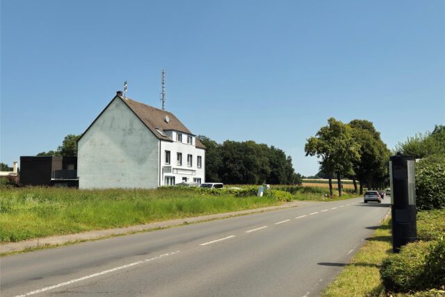 Aufnahme eines Hauses, das links im Bild neben einer Hauptverkehrsstraße in Datteln steht. Auf dem Dach des Hauses befindet sich neben einer Sirene auch ein Funkmast. Rechts im Bild eine Blitzersäule.