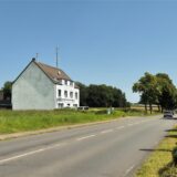 Aufnahme eines Hauses, das links im Bild neben einer Hauptverkehrsstraße in Datteln steht. Auf dem Dach des Hauses befindet sich neben einer Sirene auch ein Funkmast. Rechts im Bild eine Blitzersäule.