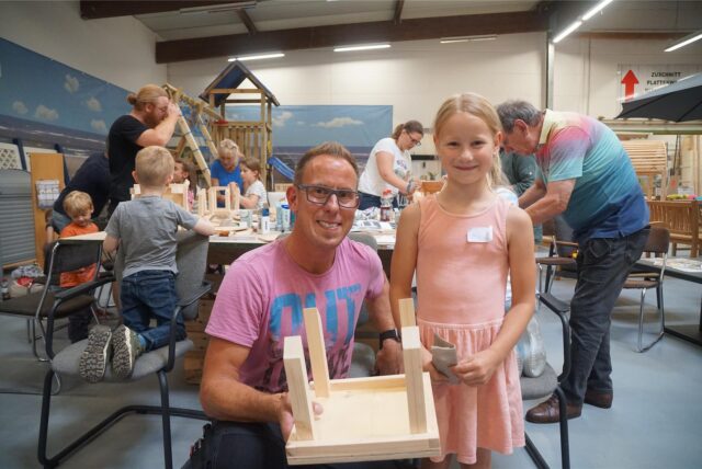 Ein Vater, mit seiner Tochter, hält ein Holzhaus in der Hand.