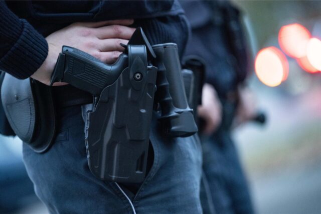 Ein Polizist steht bei einem Fototermin mit seiner Dienstwaffe an einer Straße (gestellte Szene). Nachdem er einen Schuss aus seiner Dienstwaffe bei einem Bundesligaspiel in Augsburg abgegeben haben soll, muss ein Polizist ab 20. August vor Gericht.