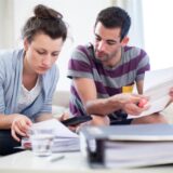 Eine Frau (links) und ein Mann sitzen auf einer Couch und studieren auf dem Tisch vor ihnen liegende Unterlagen. (Symbolbild)