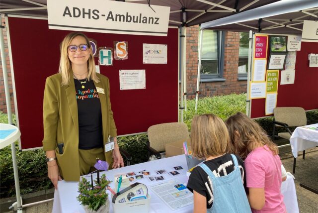 Eine Frau steht vor einer Stellwand mit der Überschrift „ADHS-Ambulanz“, vor ihr ein kleiner Tisch, vor dem zwei Mädchen stehen.
