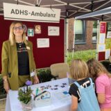 Eine Frau steht vor einer Stellwand mit der Überschrift „ADHS-Ambulanz“, vor ihr ein kleiner Tisch, vor dem zwei Mädchen stehen.