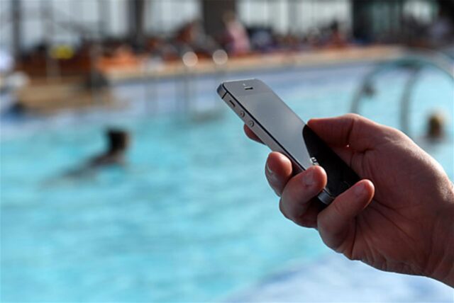 Eine Hand hält ein Smartphone, im Hintergrund ist ein Schwimmbecken zu sehen.