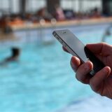 Eine Hand hält ein Smartphone, im Hintergrund ist ein Schwimmbecken zu sehen.