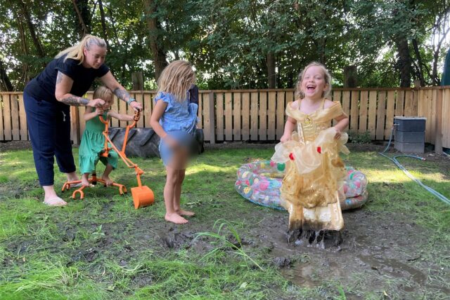 Ein Mädchen in einem langen Kleid hüpft in einer Matsch-Pfütze. Daneben steht ein anderen Mädchen, ein drittes sitzt auf einem Bagger.