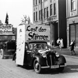 Ein historisches Foto zeigt einen Propagandazug durch Datteln. Zwei Frauen und ein Mann halten ein Plakat hoch.