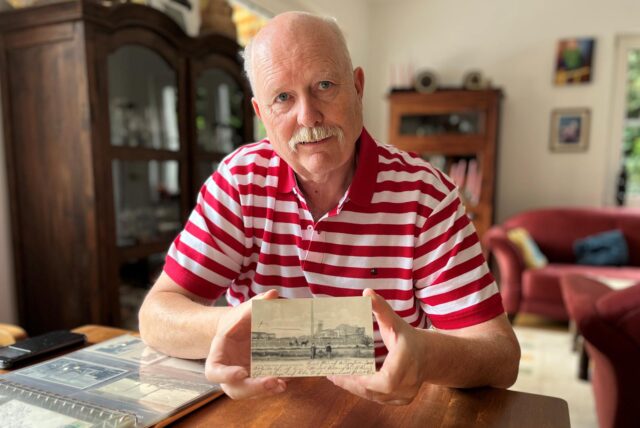 Ein Mann mit einem rot und weiß gestreiften Polohemd hält eine Postkarte in den Händen und sitzt an einem Tisch.