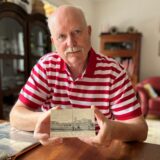 Ein Mann mit einem rot und weiß gestreiften Polohemd hält eine Postkarte in den Händen und sitzt an einem Tisch.
