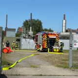 Ein Feuerwehrwagen steht auf einem Firmengelände. Feuerwehrkräfte sind im Einsatz. Wasserschläuche liegen auf dem Boden. Es ist brütend heiß, der Himmel ist blau.