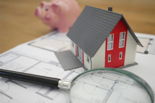 Ein Modellhaus steht auf einem Bauplan, daneben liegt eine Lupe.