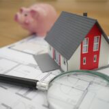 Ein Modellhaus steht auf einem Bauplan, daneben liegt eine Lupe.