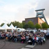Auf dem Doncaster Platz an der Zeche Ewald in Herten-Süd sitzen Männer und Frauen in mehreren Stuhlreihen. Umrahmt wird die Fläche des Sommerkinos von kleine Pavillons.
