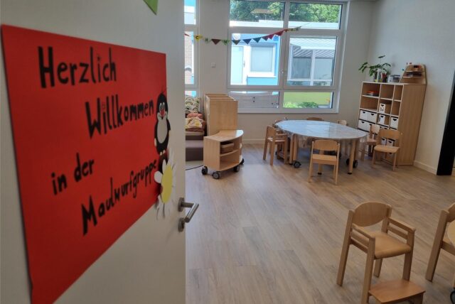 Ein Raum in einer Kita mit Mobiliar, unter anderem Stühlen und Tischen. Auf der geöffneten Tür in der linken Bildhälfte hängt ein orangefarbenes Schild, darauf die Worte „Herzlich willkommen in der Maulwurfgruppe“.
