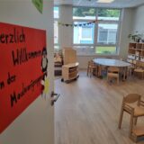Ein Raum in einer Kita mit Mobiliar, unter anderem Stühlen und Tischen. Auf der geöffneten Tür in der linken Bildhälfte hängt ein orangefarbenes Schild, darauf die Worte „Herzlich willkommen in der Maulwurfgruppe“.