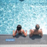 Zwei Menschen lehnen am Beckenrand eines Schwimmbads. Das Wasserreflektiert Sonnenstrahlen. Am Beckenrand ist ein Schild mit der Aufschrift „Schwimmer“ angebracht.