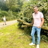 Ein Mann mit Jeans und Polohemd steht an einem umgefallenen Baum. Im Hintergrund ist ein kleiner Junge zu sehen.