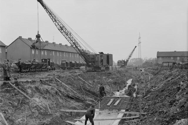 Arbeiter in einem Graben für die Kanalisierung des Herdicksbachs. Im Hintergrund ein Kran und WOhnhäuser.