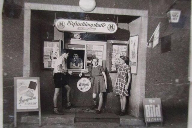 Gisela Henn (2.v.r.) im Jahr 1957 vor der „Erfrischunghalle Edith Braun“, die ihre Familie in den 50er-Jahren in der Dortmunder Nordstadt betrieb. Durch das Verkaufsfenster guckt ihre Mutter Edith Braun, links steht Gisela Henns späterer Ehemann Klaus, ganz links ein Mädchen aus der Nachbarschaft.