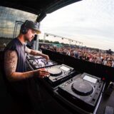 Insgesamt 1200 Gäste feierten beim Technofestival „Querfeldein“ in Recklinghausen unter Windrad in Essel mit, im Vordergrund steuert ein DJ seine Geräte.