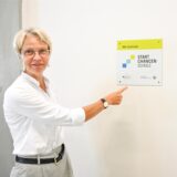 NRW Schulministerin Dorothee Feller zeigt auf die an der Wand angebrachte Plakette des „Startchancen-Programm“