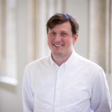 Stephan Orth steht in weißem Hemd vor einer Fensterreihe. Er lächelt.