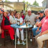 Eine Gruppe von Menschen steht um einen Stehrisch in einer Fußgängerzone. Auf dem Tisch stehen Bierflaschen und Weingläser. Die Personen tragen sommerliche Kleidung. Im Hintergrund ist ein Bierwagen zu erkennen.