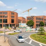 Die Baustelle der Hertener Höfe. Rechts davor ist der Kreisverkehr in Herten-Mitte zu sehen, durch den ein weißes Auto fährt.