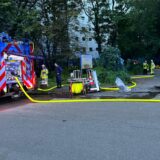 Ein Feuerwehrwagen und Einsatzkräfte stehen vor einem großen Hochhaus. Vor dem Haus stehen Bäume und Büsche, die den Blick verwehren.