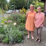 Barbara Schweda und Bettina Marquard (r.) stehen neben bunten Blumen in einem Vorgarten.