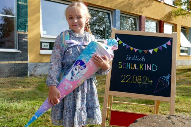 Anne Sophie seht an der Süder Grundschule in Herten neben einer Tafel, auf dem steht, dass sie endlich Schulkind ist, versehen mit dem Datum ihrer Einschulung.