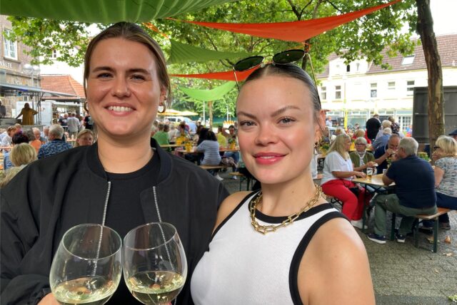 Zwei junge Frauen stehen auf einem Platz und halten mit Weißwein gefüllte Gläser in den Händen.