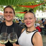 Zwei junge Frauen stehen auf einem Platz und halten mit Weißwein gefüllte Gläser in den Händen.