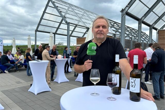 Redakteur Ralf Wiethaup steht mit Mikrofon an einem Bistrotisch im Freien, auf dem zwei Flaschen Wein stehen und zwei gefüllte Weingläser.