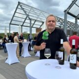 Redakteur Ralf Wiethaup steht mit Mikrofon an einem Bistrotisch im Freien, auf dem zwei Flaschen Wein stehen und zwei gefüllte Weingläser.