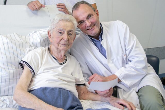 Ein älterer Patient und ein Chefarzt im weißen Kittel sitzen auf einem Krankenbett und lächeln in die Kamera.