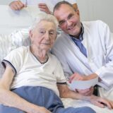 Ein älterer Patient und ein Chefarzt im weißen Kittel sitzen auf einem Krankenbett und lächeln in die Kamera.