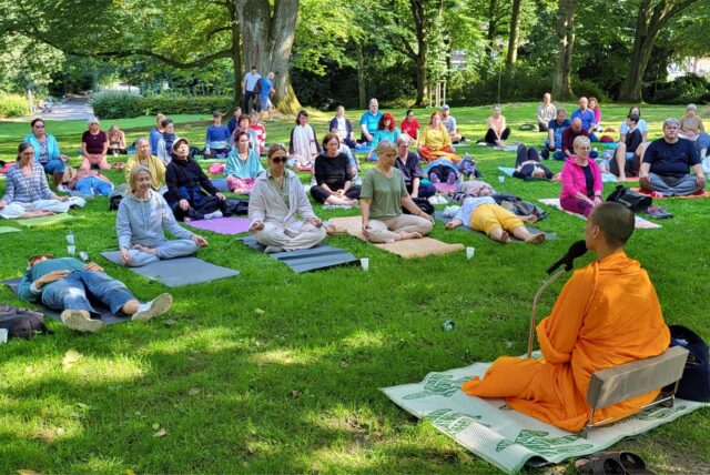 Der buddhistische Mönch Phra Puttipong aus Moers sitzt mit Mikrofon in der Hand im Stadtgarten Recklinghausen im Schneidersitz vor rund 100 meditierenden Menschen.