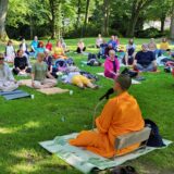 Der buddhistische Mönch Phra Puttipong aus Moers sitzt mit Mikrofon in der Hand im Stadtgarten Recklinghausen im Schneidersitz vor rund 100 meditierenden Menschen.