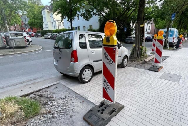 Ein Auto steht zwischen zwei Bäumen. Zwei Schilder sind zu sehen, die noch auf eine Baustelle hindeuten.