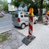 Ein Auto steht zwischen zwei Bäumen. Zwei Schilder sind zu sehen, die noch auf eine Baustelle hindeuten.