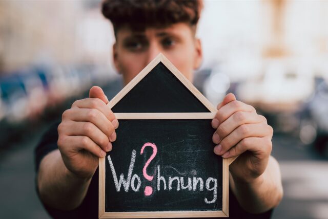 „Wo?hnung“ steht in Kreideschrift auf einer hausförmigen Tafel, die ein junger Mann mit beiden Händen in die Kamera hält.
