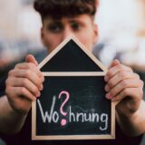 „Wo?hnung“ steht in Kreideschrift auf einer hausförmigen Tafel, die ein junger Mann mit beiden Händen in die Kamera hält.