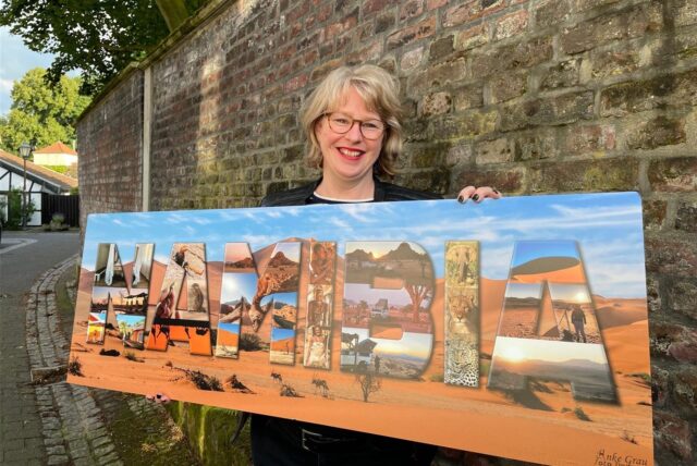 Anke Grau steht mit einem großen Fotoplakat vor der Mauer auf der Freiheit in Westerholt. Auf dem Bild ist eine Dünenlandschaft einer Wüste zu erkennen, davor steht in Großbuchtstaben „Namibia“ wobei die Buchstaben mit vielen Motiven wie Tieren der Savanne oder weiteren Wüstenmotiven gefüllt sind.