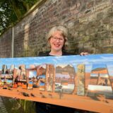 Anke Grau steht mit einem großen Fotoplakat vor der Mauer auf der Freiheit in Westerholt. Auf dem Bild ist eine Dünenlandschaft einer Wüste zu erkennen, davor steht in Großbuchtstaben „Namibia“ wobei die Buchstaben mit vielen Motiven wie Tieren der Savanne oder weiteren Wüstenmotiven gefüllt sind.